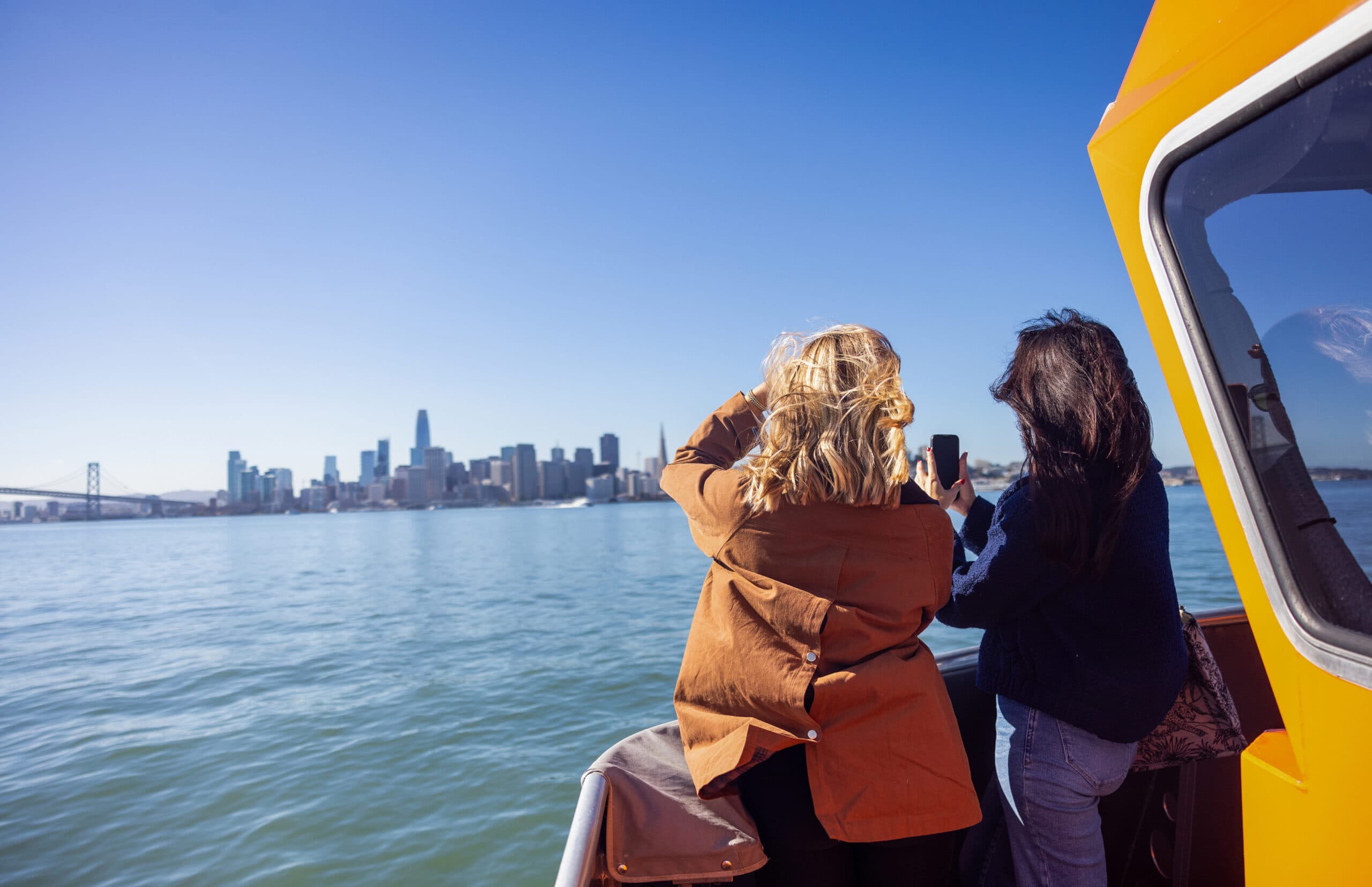 Two women standing on a ferry deck photographing the San Francisco skyline and Bay Bridge across calm bay waters on a bright, clear day.