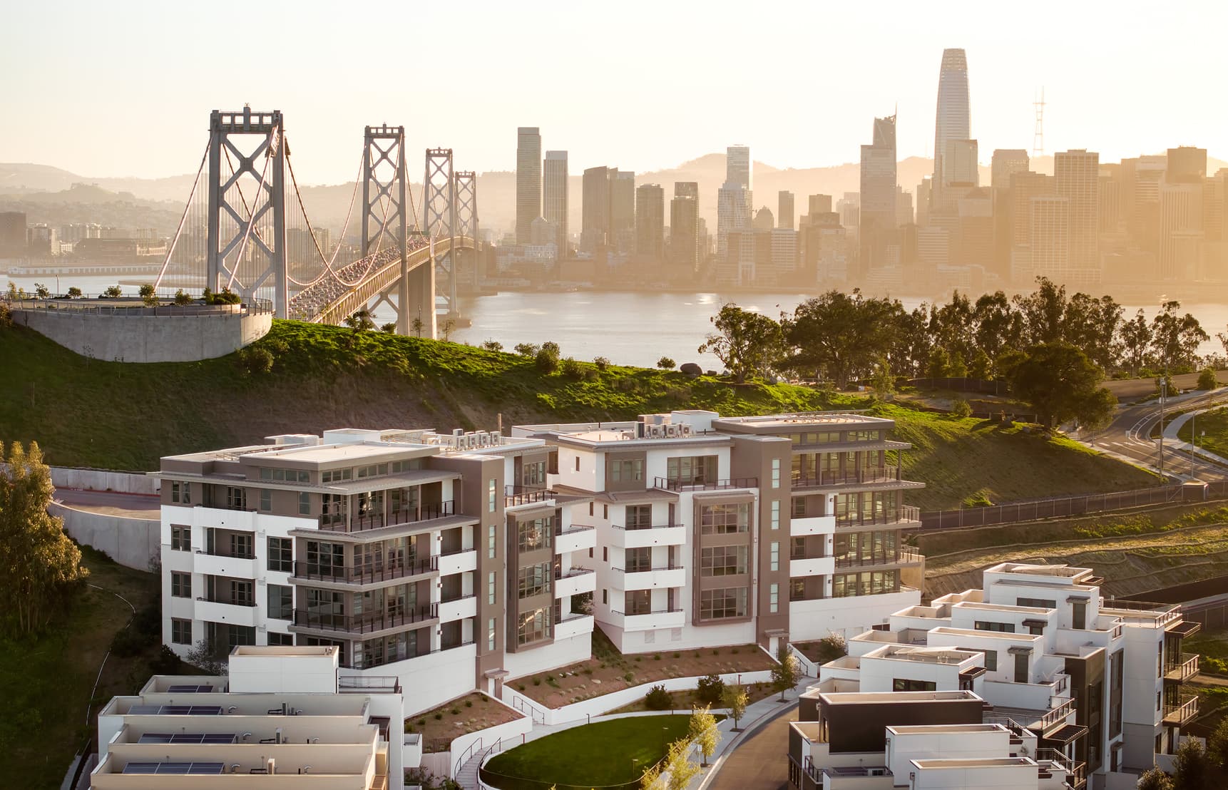 Modern condominium buildings at The Flats on Treasure Island overlooking the San Francisco Bay, with the Bay Bridge and downtown skyline glowing at sunset in the background.