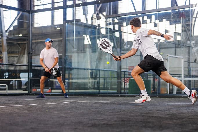 Two men playing padel inside a glass-walled court, with one player lunging forward to return a low ball while his partner stands ready behind him.