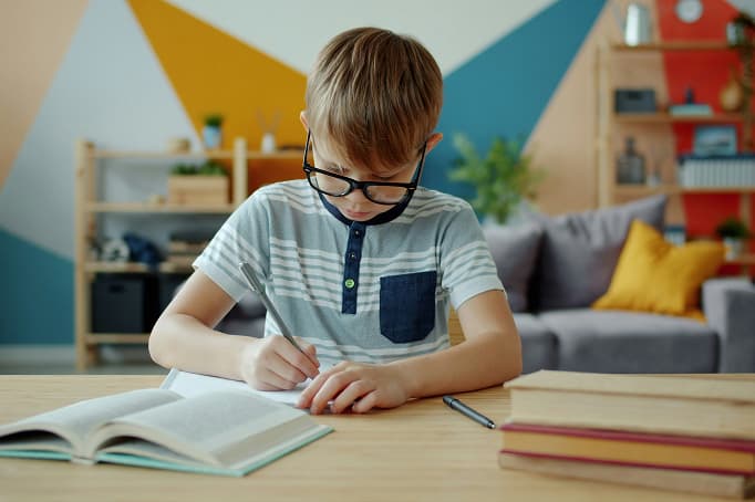 A young boy wearing glasses sits at a table, concentrating as he writes in a workbook, with books stacked nearby in a colorful home setting.