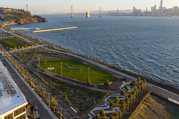 An aerial view of a waterfront park with walking paths, landscaped greenery, and a large open lawn overlooking the bay, with the Bay Bridge and San Francisco skyline visible in the distance.