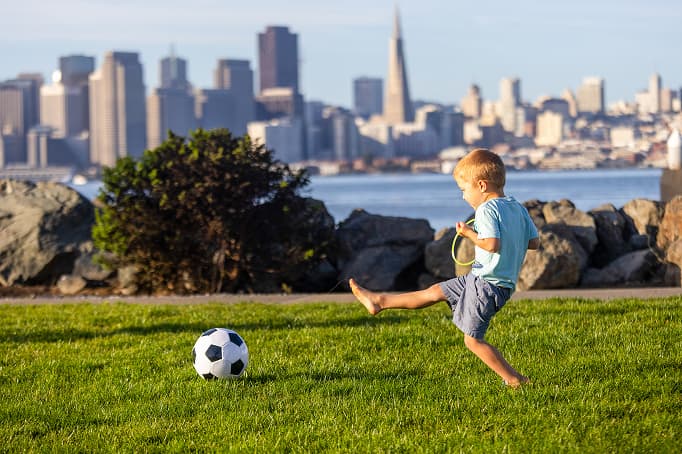 A young child kicking a soccer ball on a grassy waterfront park, with the San Francisco skyline and Transamerica Pyramid visible across the bay in the background.