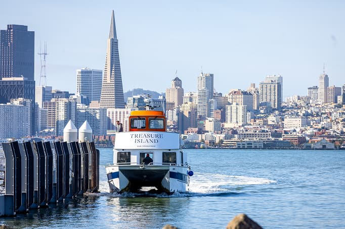 A ferry labeled “Treasure Island – San Francisco” approaches a dock, with the San Francisco skyline and the Transamerica Pyramid rising in the background across the bay.