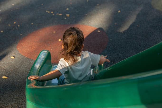 A young child sliding down a green playground slide, seen from behind, with sunlight casting soft shadows across the rubber play surface below.