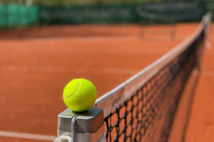 A bright yellow tennis ball resting on the metal post of a net on a clay tennis court, with the net and court surface stretching out of focus in the background.