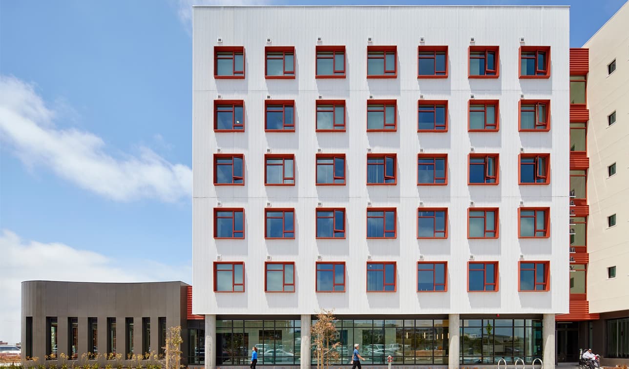 Exterior of the Maceo May apartment building with a white facade, red-trimmed windows, and people walking past the ground-floor entrance on a clear day.