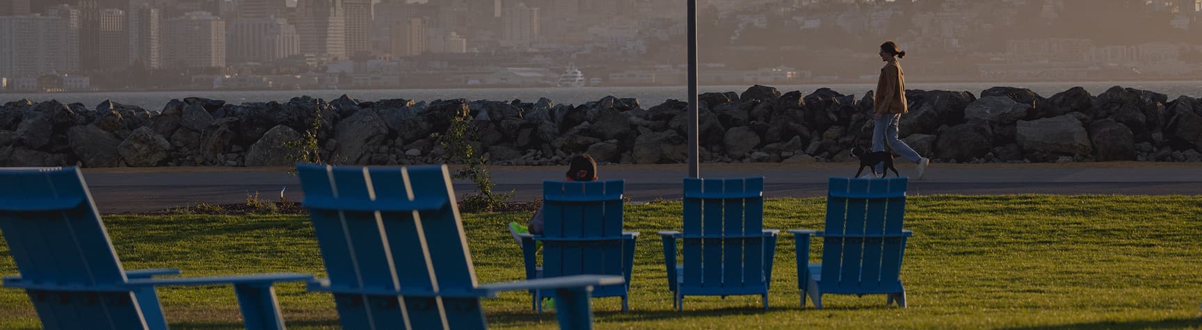Waterfront view of Treasure Island with open green space and pathways in the foreground, overlooking San Francisco Bay with the city skyline and Bay Bridge visible in the distance.
