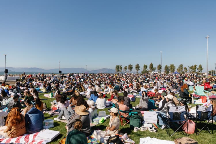 Large crowd gathered on a waterfront park lawn for a sunny outdoor community festival with picnic blankets, lawn chairs, and boxed food.