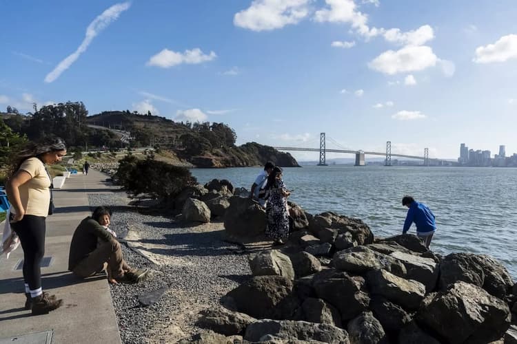 People relaxing and exploring along a rocky waterfront trail with the San Francisco skyline and Bay Bridge visible across the water on a clear day.