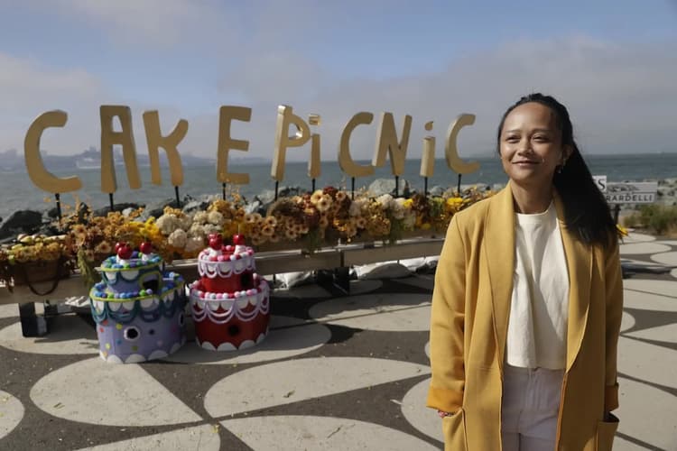 A woman stands smiling at a waterfront celebration setup with large gold letters spelling “CAKE PICNIC” behind her, decorated with flowers and colorful cake displays.