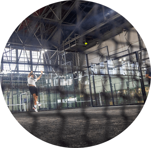 Indoor padel player jumping to serve on a modern glass court inside an industrial-style sports facility with metal beams and high ceilings, showcasing competitive racket sports training.