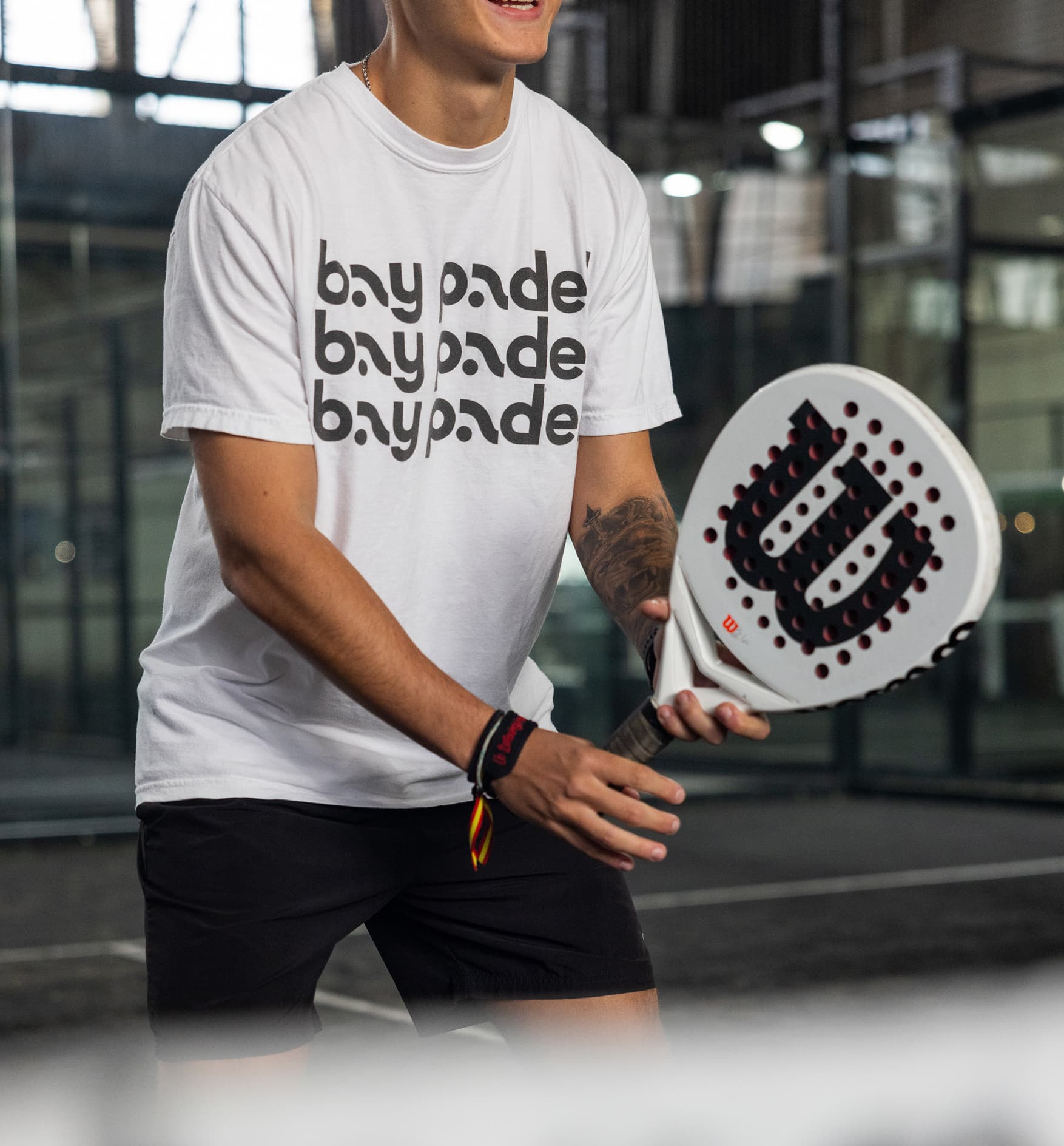 Close-up of a padel player wearing a Bay Padel T-shirt and holding a Wilson padel racket on an indoor glass court, highlighting branded athletic apparel and competitive racket sports.