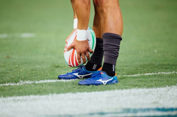 A close-up of an athlete standing on a grass field, holding a rugby ball near the sideline, wearing blue cleats and black socks, preparing to take a kick or restart play.
