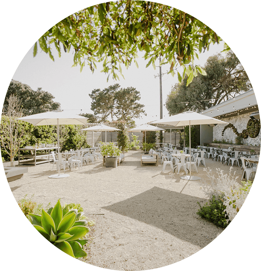 Sunny outdoor patio at Aracely Café on Treasure Island featuring garden landscaping, shaded umbrellas, communal tables, and mural art for al fresco dining in San Francisco.