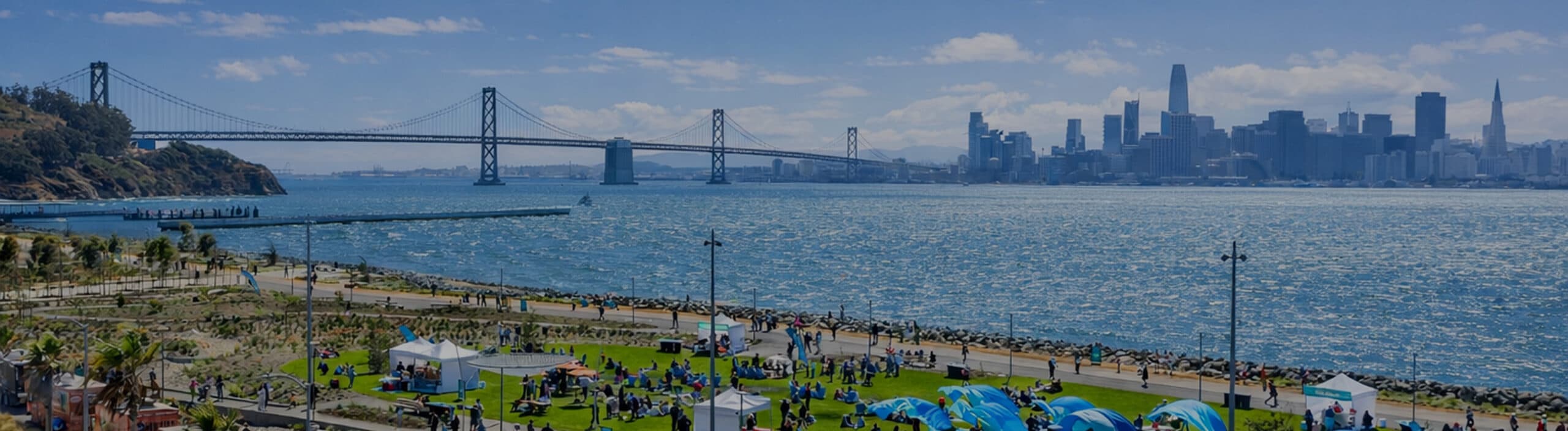 People enjoying an outdoor event at Cityside Park on Treasure Island with blue tents, food trucks, and views of the Bay Bridge and San Francisco skyline.